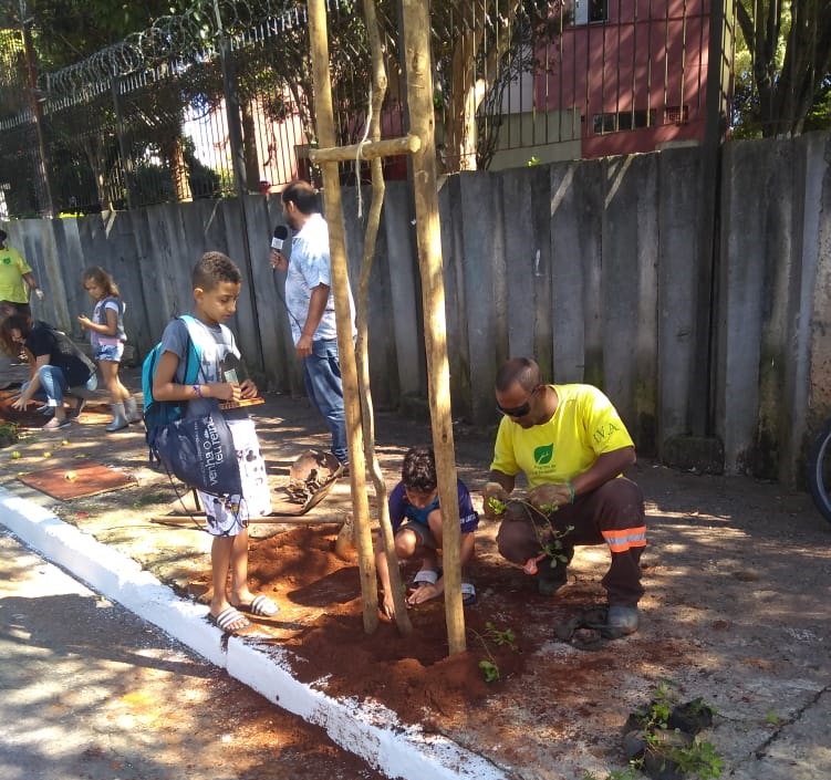 A imagem mostra um homem de camiseta amarela junto de dois garotos com camisetas azuis, de aproximadamente sete anos cada um. Os três estão plantando uma muda de árvore na calçada da rua Barão do Rio da Prata.